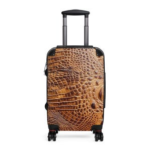 May include: A rolling suitcase featuring a brown and tan textured crocodile skin pattern on the front panel. The luggage has a black frame, a retractable silver metal handle, and four black spinner wheels at the base for mobility.