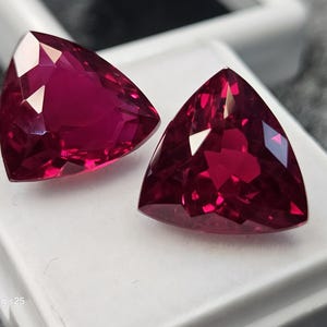 以下が含まれることがあります： 深い鮮やかなマゼンタレッドの色合いを持つ、トリリアントカットのルースストーン2石。これらのファセットカットされた石は、鋭い先端を持つ三角形で、反射する表面を備えています。Samsung s25というテキストが記された白い台座の上に置かれています。