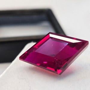 May include: A square-cut, vibrant magenta gemstone with faceted surfaces that reflect light. The stone is positioned at an angle, showcasing its sharp edges and deep, saturated pinkish-red color, with a dark display box visible in the background.