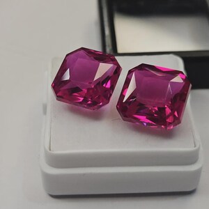 May include: Two vibrant, cushion-cut fuchsia gemstones with faceted surfaces that reflect light. The stones are positioned side-by-side on a white display pad, which is part of a small, open black jewelry box with a white interior lining.