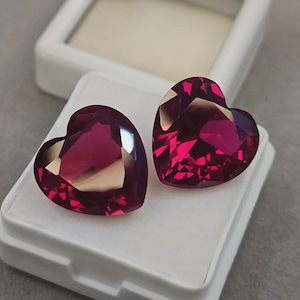 May include: Two faceted heart-shaped gemstones in a deep magenta-red colour. The stones feature precise geometric cuts that create light reflections across their surfaces. They are positioned side-by-side on a white rectangular display base.