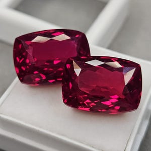 Peut inclure: Deux pierres précieuses rouge profond de taille coussin avec des surfaces facettées qui reflètent la lumière. Les pierres sont placées sur une base de présentation blanche. Le mot surbhit est imprimé dans le coin inférieur gauche.