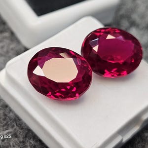 May include: Two oval-cut, deep red gemstones with faceted surfaces, displayed on a white square display box. The stones exhibit a vibrant, translucent crimson hue with sharp geometric reflections across their polished tops.