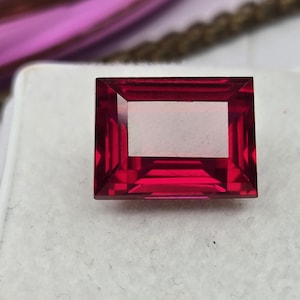 May include: A rectangular, faceted gemstone with a deep, vibrant red color. The stone features a step-cut design with clear, geometric lines and a polished surface that reflects light, showcasing its transparent and saturated crimson hue.