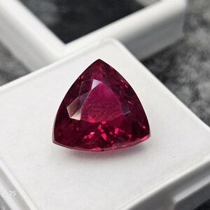 May include: A deep magenta-red gemstone cut into a triangular trillion shape with multiple facets that reflect light. The gemstone is displayed resting on a white square display box with a clear lid, featuring the text Samsung s25 in the corner.