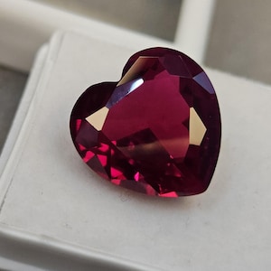 May include: A faceted heart-shaped gemstone with a deep, vibrant magenta-red color. The stone features multiple triangular and trapezoidal facets that reflect light, creating sharp highlights and deep shadows across its polished, translucent surface.
