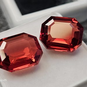 May include: Two loose, rectangular, emerald-cut gemstones with a vibrant, translucent red hue. The stones feature faceted surfaces that reflect light, creating depth and clarity. They are positioned side-by-side on a white display surface.