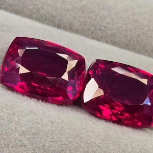 Può includere: Due pietre preziose sciolte con taglio a cuscino e un colore rosso magenta profondo e vibrante. Ogni pietra presenta una superficie sfaccettata che riflette la luce, creando nitidi motivi geometrici sulla parte superiore. Le pietre sono affiancate.