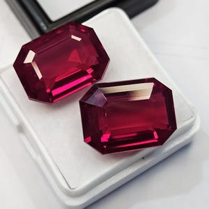May include: Two deep red, emerald-cut gemstones with faceted surfaces, displayed on a white rectangular jewellery display pad. The stones exhibit a rich, saturated crimson hue with reflective highlights across their polished, geometric edges.