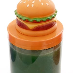 May include: A cylindrical clear plastic container filled with a dark green substance, topped with an orange lid and a decorative burger-shaped topper featuring a tan bun with white sesame seeds, green lettuce, yellow cheese, and a red patty.