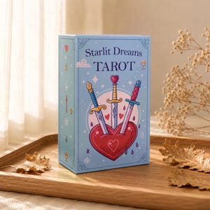 May include: A light blue rectangular tarot deck box titled Starlit Dreams TAROT. The front features a large red heart pierced by three decorative swords, surrounded by small stars and sparkles. The side panel displays various small icons.