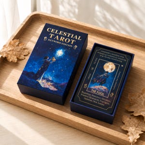 May include: A Celestial Tarot Keyword Edition deck in a dark blue box with gold lettering. The box lid features a figure reaching toward a glowing star. Inside the open box, a card titled The Fool displays a figure under a full moon.