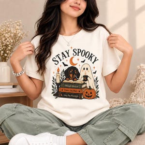 May include: A cream-colored short-sleeve t-shirt featuring a graphic print with the text STAY SPOOKY, a dachshund, a ghost holding a mug, a stack of books titled Midnight Mischief, Wiener Dog Magic, and Long Dog Moonlight, a candle, and a pumpkin.