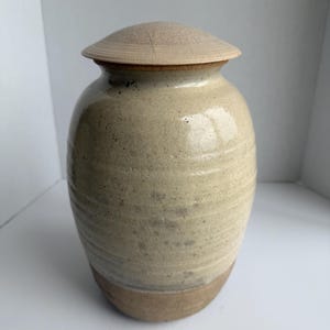 May include: A ceramic jar with a speckled cream-coloured glaze and a rounded body featuring horizontal ridges. The jar is topped with a matching, slightly domed, circular lid that has a textured, matte finish. The base is unglazed and brown.