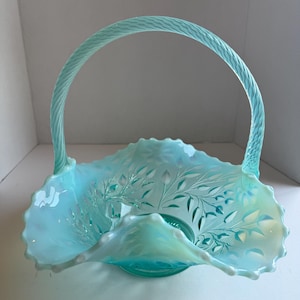 May include: A light blue opalescent glass basket featuring a ruffled, scalloped rim and a textured, woven-style handle. The body of the basket is decorated with an intricate, embossed floral and leaf pattern across the interior surface.
