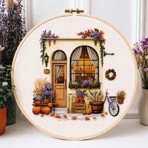 Puede incluir: Un bastidor de bordado circular con un diseño en punto de cruz de la fachada de un edificio con puerta y ventana. La escena incluye calabazas naranjas, flores moradas, enredaderas verdes y una bicicleta, bordados sobre tela blanca.