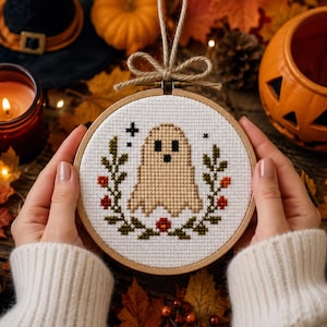May include: A circular wooden embroidery hoop containing a cross-stitch design on white fabric. The design features a tan ghost with black eyes and a mouth, surrounded by a wreath of green leaves and small red berries, with two black stars.