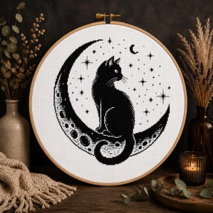 Peut inclure: Un tambour à broder circulaire en bois contenant un motif au point de croix sur tissu blanc. Le motif représente un chat noir assis sur un croissant de lune, entouré de plusieurs petites étoiles et d'un petit croissant de lune en fil noir.