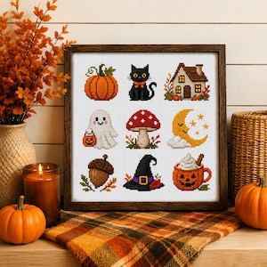 May include: A framed cross-stitch sampler featuring nine distinct autumn-themed icons arranged in a grid: an orange pumpkin, a black cat, a house, a ghost holding a jack-o'-lantern, a red mushroom, a crescent moon, an acorn, a witch hat, and a mug.