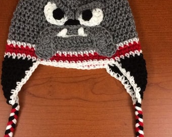 May include: A handmade crochet hat in the shape of a bulldog. The hat is primarily gray with white, black, and red accents. It features a bulldog face with large eyes and floppy ears. Braided tassels hang from the ear flaps.