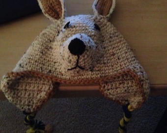 May include: A crocheted hat shaped like a kangaroo with brown ears, a white face, and black eyes. The hat has yellow and black yarn tails.