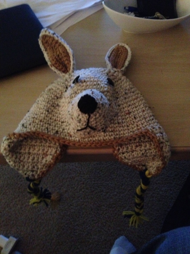 May include: A crocheted hat shaped like a kangaroo with brown ears, a white face, and black eyes. The hat has yellow and black yarn tails.