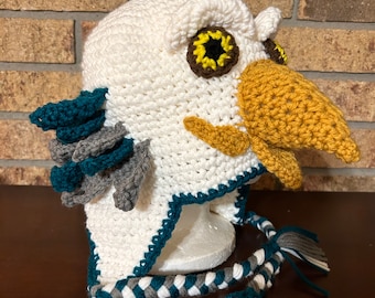 May include: A handmade white crochet owl hat with yellow, brown, teal, and gray accents. The hat features large owl eyes, a beak, and feather details. Braided ties with tassels complete the design.