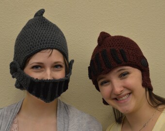 May include: Two people wearing crocheted hats shaped like helmets. The hats are gray and burgundy with black accents. The person on the left is wearing a gray hat with a black face shield. The person on the right is wearing a burgundy hat with black trim.