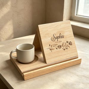May include: A light-colored wooden stand with a triangular easel design featuring the engraved name Sophia, the date 11.1.2009, and a floral branch illustration. A plain off-white ceramic mug sits in a circular cutout on the base of the stand.