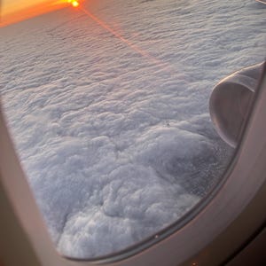 May include: A view from an airplane window showing a bright orange and yellow sunset over a vast layer of white clouds. The airplane wing and engine are visible on the right side of the frame, with a lens flare extending from the sun.