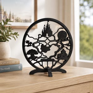 May include: A black circular decorative stand featuring a laser-cut silhouette map of a theme park. The design includes iconic structures such as a central castle, a geodesic dome, a large tree, and palm trees, all connected by winding pathways.
