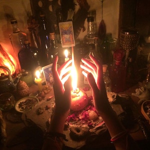 May include: A bright orange candle flame burns between two hands, positioned above a stone surface scattered with dried flower petals, small stones, and a tarot card. Several glass bottles and a small lit candle are visible in the background.