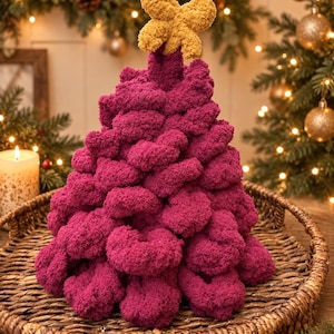 Op de afbeelding: Een kegelvormige kerstboomdecoratie gemaakt van dikke, getextureerde magenta garenlussen. De boom heeft een opvallende, mosterdgele, vierbladige bloemvormige piek. De gehele structuur is vervaardigd uit zacht, pluche materiaal.