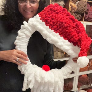 May include: A circular wreath crafted from chunky white and red yarn, designed to resemble a Santa Claus face with a red hat, a white brim, a white beard, and a small red pom-pom nose. The wreath features a soft, textured finish.