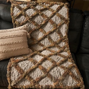 May include: A chunky knit throw blanket featuring a cream-colored base with a raised, diamond-shaped lattice pattern in variegated shades of brown, tan, and green. A rectangular cream-colored throw pillow with horizontal textured stripes sits nearby.