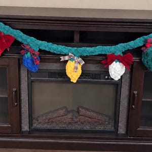 May include: A long, teal-colored crocheted garland featuring plush, bulb-shaped ornaments in white, yellow, red, and blue. Each bulb is accented with a small fabric bow in plaid or solid red, creating a decorative string of holiday-themed lights.