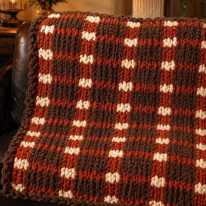 May include: A chunky knit throw blanket featuring a plaid pattern in dark brown, burnt orange, and cream. The blanket is constructed from thick, soft yarn loops, creating a textured surface with a consistent grid design throughout the fabric.
