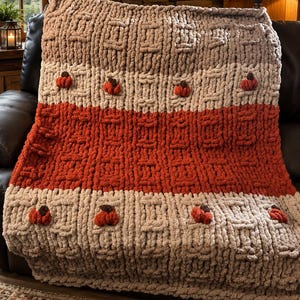 May include: A chunky knit throw blanket featuring horizontal stripes in shades of tan, cream, and burnt orange. The blanket has a textured basketweave pattern and is decorated with small, plush, orange pumpkin-shaped appliques with brown stems.