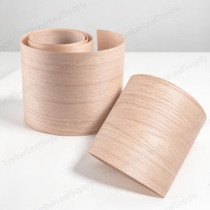 May include: Two rolls of light-coloured wood veneer edge banding featuring a natural grain pattern. The material is flexible and thin, showing a smooth, light tan surface with horizontal wood grain lines visible across the curved surfaces.