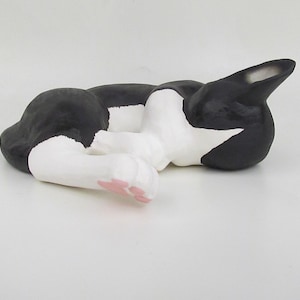 May include: A ceramic figurine of a sleeping cat in a curled position. The cat has a black and white coat with distinct white paws and a small pink detail on the underside of one paw. The ears are pointed and black, and the eyes are closed.