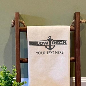 May include: A white kitchen towel with a black anchor and the text "BELOW DECK" and "YOUR TEXT HERE". The towel is hanging on a wooden ladder.