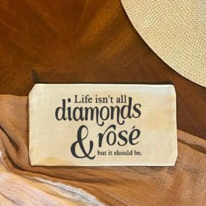 May include: A rectangular off-white canvas zipper pouch featuring the black printed text Life isn't all diamonds & rosé but it should be. The pouch has a black zipper closure along the top edge and a simple, flat, structured design for storage.