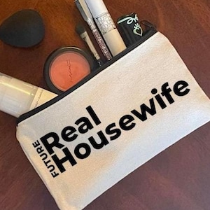May include: White canvas makeup bag with a black zipper and the text "FUTURE REAL HOUSEWIFE" printed in black.