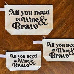 May include: Three canvas zipper pouches with the text "All you need is Wine & Bravo" in black. The pouches are off-white with a white strap and a silver zipper. The pouches are different sizes.