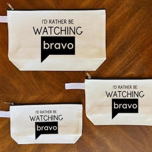 May include: Three off-white canvas zipper pouches with the text "I'D RATHER BE WATCHING bravo" in black. The pouches vary in size and have a white fabric pull tab attached to the zipper.