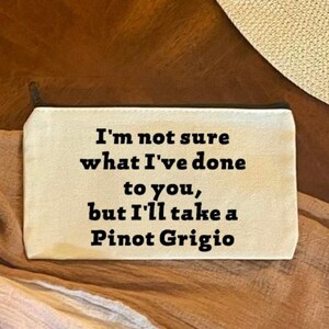 May include: A beige canvas pouch with the text "I'm not sure what I've done to you, but I'll take a Pinot Grigio" in black. The pouch is on a brown surface with a straw hat in the background.