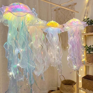 May include: A set of four hanging jellyfish-shaped decorations made from iridescent, translucent fabric. Each piece features a rounded bell top and long, flowing, wavy tentacles illuminated by internal string lights that emit a soft glow.