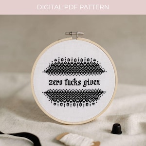 May include: A circular wooden embroidery hoop containing white fabric with a black cross-stitch design. The design features decorative geometric borders above and below the text "zero fucks given" stitched in a bold, black, sans-serif font.