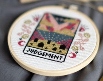 Może przedstawiać: Tamborek do haftu krzyżykowego z kolorowym wzorem karty tarota ze słowem "JUDGEMENT" na dole. Projekt zawiera kwiatowe i półksiężycowe akcenty, na białym tle w drewnianym tamborku. Tekst "DIGITAL PDF PATTERN" znajduje się na górze.