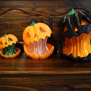 May include: Three jack-o'-lantern decorations in varying sizes. The smallest is orange with a green interior, the middle is orange with a clear interior, and the largest is black with an orange interior. Each features a carved face design.